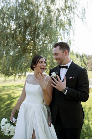 A joyful wedding couple shares a candid moment together under a willow tree with white floral bouquet.