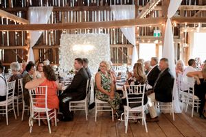 Reception guests seated at decorated tables inside rustic barn wedding venue with twinkling lights and white drapes.