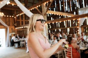 A guest wearing sunglasses and a pink top laughs joyfully during a celebration inside a rustic barn venue with string lights.