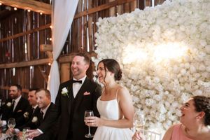 Wedding celebration at a rustic barn venue with elegant white floral backdrop and guests enjoying dinner reception.