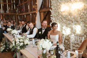 Wedding party seated at an elegant rustic barn reception with white floral decor and warm lighting.