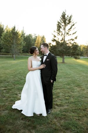 A bride in white gown and groom in black tuxedo embrace on grassy lawn with pine trees in background.