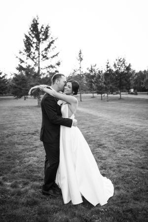 A couple embraces outdoors on their wedding day in a black and white photograph taken in a grassy field with trees.