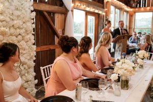 Wedding reception guests seated at a rustic farmhouse table with white decor inside a barn venue.