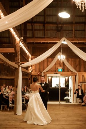 A couple shares their first dance in a rustic barn venue decorated with white drapes and twinkling lights.
