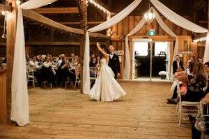 A couple shares their first dance in a rustic barn reception venue decorated with white drapes and string lights.