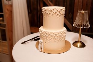 A two-tier white wedding cake decorated with cascading pearl-like details sits on a round table with a small table lamp.