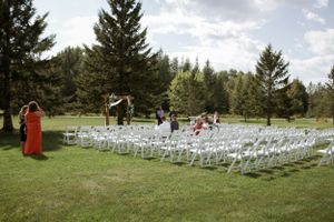 Rows of white chairs set up for an outdoor wedding ceremony on a grassy lawn surrounded by tall evergreen trees.