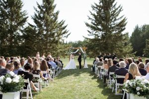 Outdoor wedding ceremony on a grassy field with white chairs and evergreen trees creating a natural backdrop.