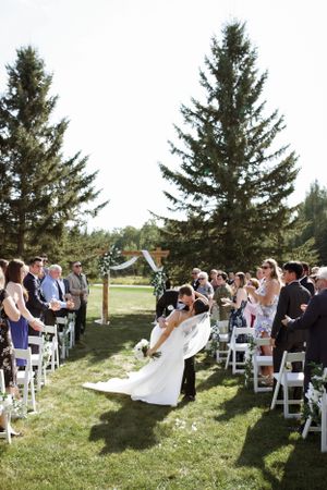 Outdoor wedding ceremony setup with white chairs on a lawn surrounded by tall evergreen trees in the background.