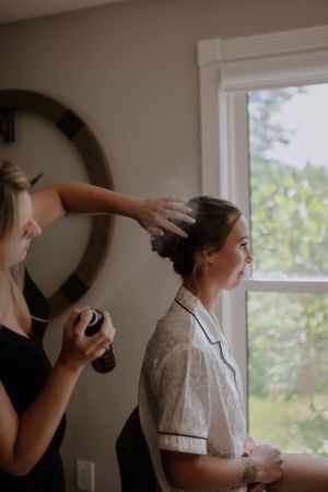 Hair stylist applies product and styling to someone's hair in a sunlit room with a circular mirror on the wall.
