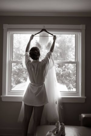 Black and white photo of someone in a robe hanging up a wedding dress by a sunlit window with ballet slippers below.