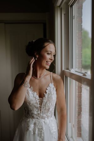 A bride in a delicate lace wedding gown with spaghetti straps stands by a window in natural light.