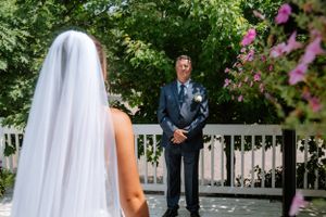 A wedding couple during their first look moment outdoors with pink flowers and a white fence in the background.