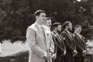Row of groomsmen in suits stand together outdoors during wedding ceremony.