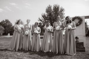 A line of bridesmaids in flowing gray dresses stand together outdoors in a black and white wedding photograph.