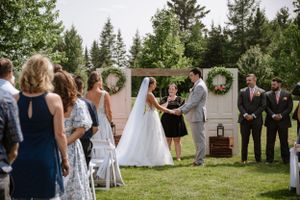 Outdoor wedding ceremony with rustic backdrop, decorative wreaths and gathered guests on lawn.