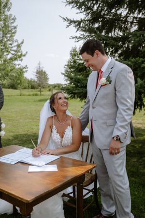 Happy couple at outdoor wedding ceremony signing marriage license at wooden table near evergreen trees.