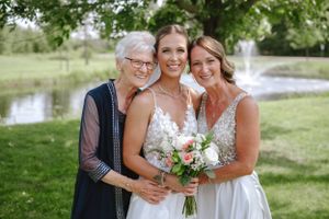 A joyful wedding photo showing three women in elegant dresses embracing outdoors by a pond on a sunny day.