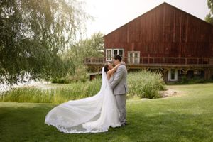Wedding couple embraces on green lawn in front of rustic red barn with willow tree on sunny summer day.
