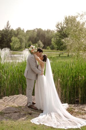 A couple embraces by a pond with tall grass, the bride's flowing white dress trailing behind her in a natural outdoor setting.