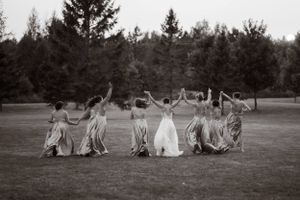 Black and white photo of bridesmaids and bride sitting in a circle on grass with raised arms during wedding celebration.