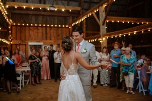 A couple shares their first dance in a rustic barn venue decorated with string lights as guests look on.