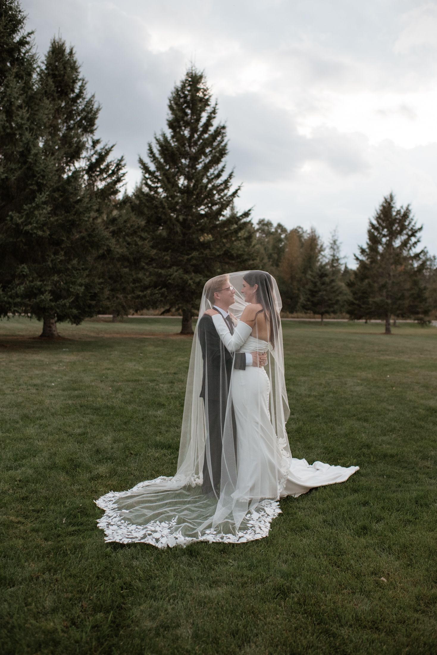 A romantic wedding moment captured under evergreen trees with a flowing lace veil.