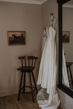 An elegant white wedding dress hangs in a room with wooden bar stools and framed horse artwork on neutral walls.
