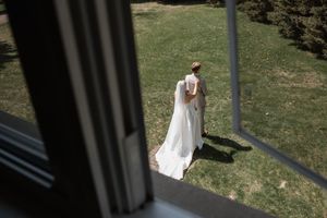 A couple in elegant wedding attire walks through a sunny garden with lush greenery in the background.