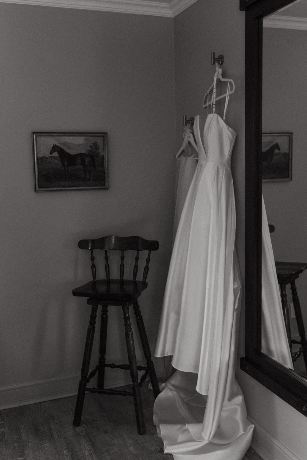 A white wedding dress hangs next to a wooden bar stool in a room with horse paintings on the wall, captured in black and white.