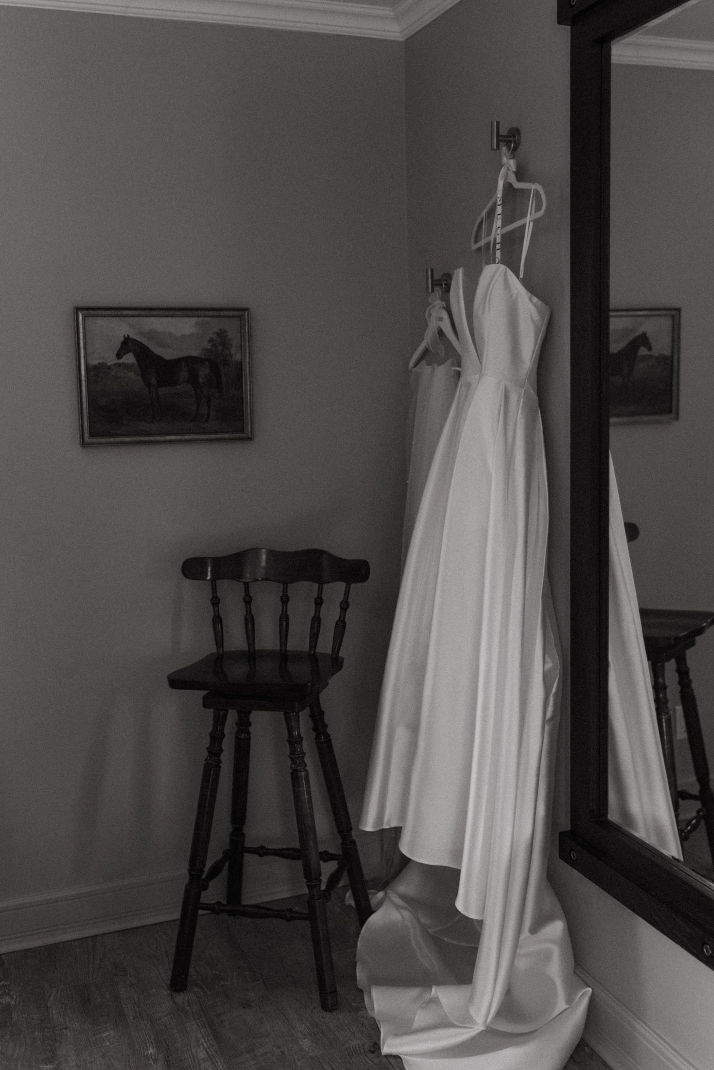 A white wedding dress hangs next to a wooden bar stool in a room with horse paintings on the wall, captured in black and white.