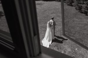Black and white photo taken through window showing couple in formal attire walking on grass in sunlight.