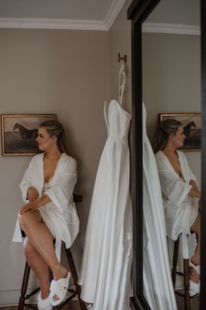 A wedding dress hangs near a mirror while preparing with white slippers and horse artwork on the wall.