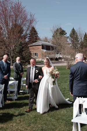 An outdoor spring wedding ceremony taking place on a grassy lawn with white chairs and evergreen trees in the background.