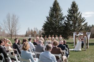 Outdoor wedding ceremony in progress with guests seated on white chairs facing wooden arch.