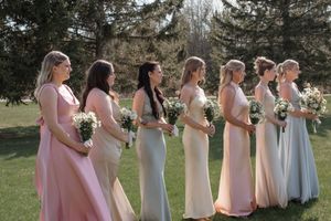 Seven bridesmaids in pastel dresses stand in a row outdoors holding bouquets during a spring wedding ceremony.