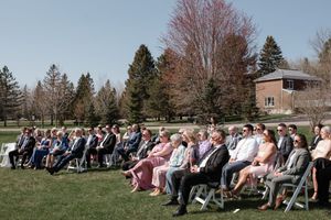 Wedding guests seated in rows of white chairs on a grassy lawn during an outdoor ceremony with evergreen trees in background.