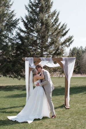 Couple sharing first kiss under white draped arch with evergreen trees in background.