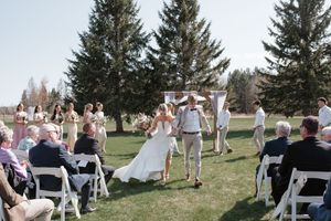 Wedding party celebrates on grassy hillside with trees in background during outdoor ceremony.