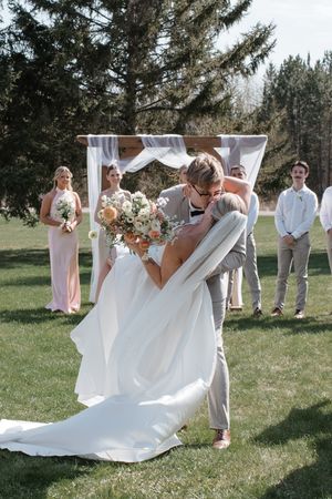 Couple shares romantic kiss under white fabric arch during outdoor wedding ceremony with wedding party and pine trees in background.