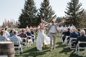 A panoramic view of an outdoor wedding ceremony with guests seated on white chairs surrounded by evergreen trees.