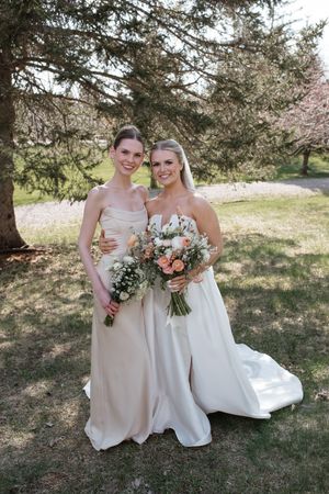 Two women wearing formal gowns stand together under a pine tree holding floral bouquets during a wedding celebration.