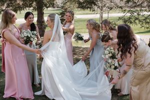 Bridesmaids in pastel dresses help arrange the bride's flowing white veil and gown in an outdoor garden setting.