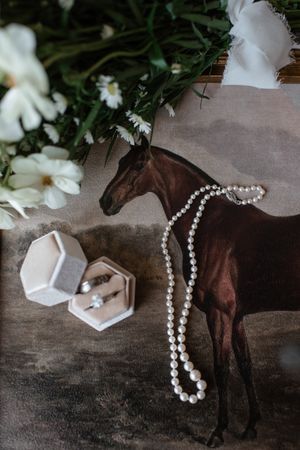 Wedding details flatlay with horse figurine, pearl necklace, ring box and white flowers on grey background.