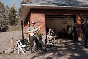 Musicians perform outside a brick garage during a casual outdoor gathering in a wooded setting.