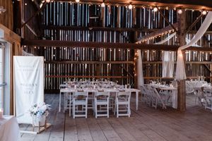 Rustic barn wedding reception setup with white tables and chairs under string lights and wooden beams.