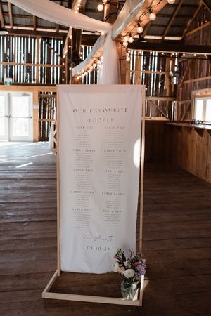 White fabric seating chart display in a rustic barn venue with twinkle lights and draped fabric decorations.