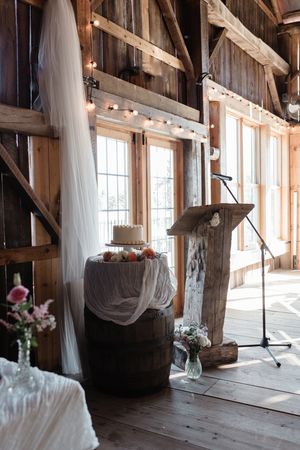 Rustic barn wedding setup with wooden podium, delicate white drapes, and cake displayed on wine barrel near window.