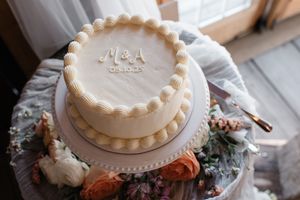 White two-tiered wedding cake with monogram initials and pearl border details on decorative white cake stand.